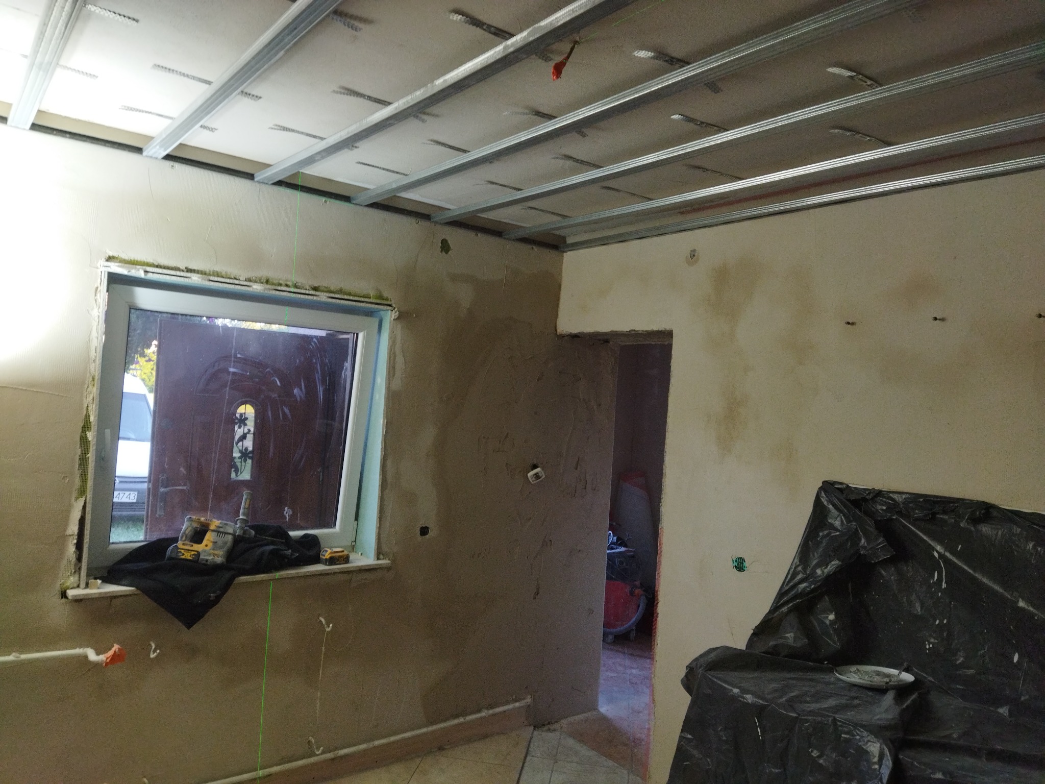 Wnętrze w trakcie remontu: widoczne metalowe profile sufitowe, częściowo otynkowane ściany, okno z narzędziami na parapecie i zabezpieczona folią część pomieszczenia. W tle widoczny odkurzacz...