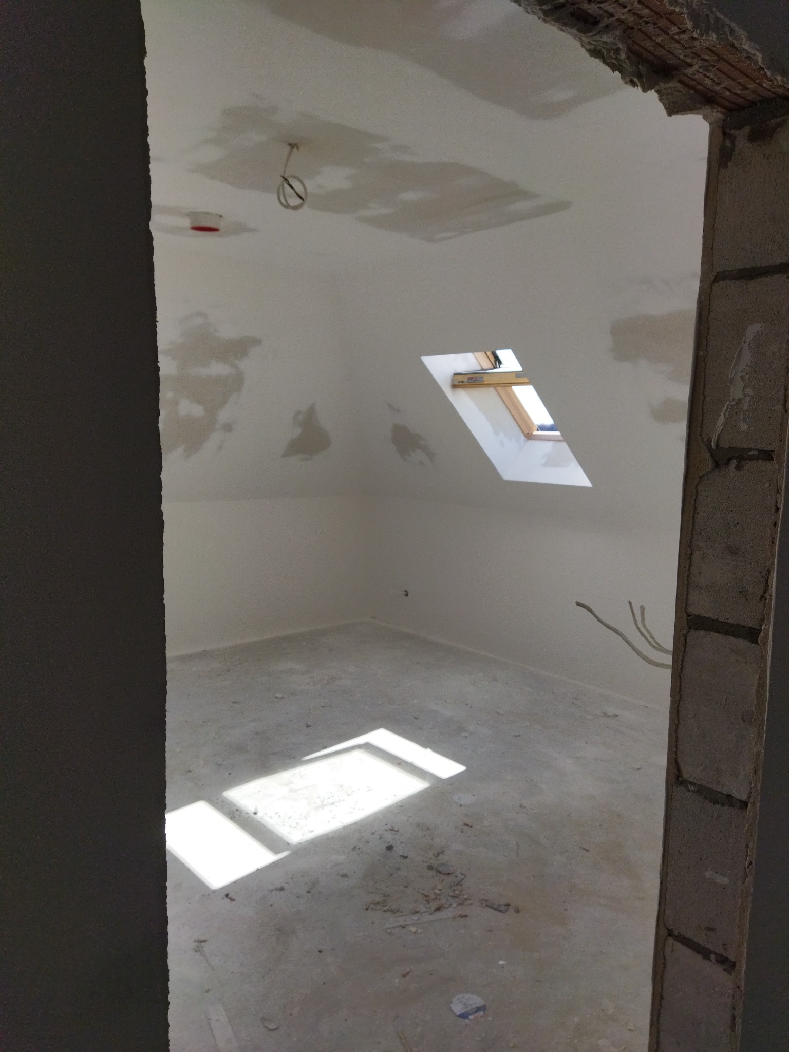 Poddasze w trakcie remontu: widok na surowe ściany z widocznymi śladami szpachlowania, okno dachowe wpuszczające światło na betonową podłogę, wystające przewody elektryczne i niedokończone...