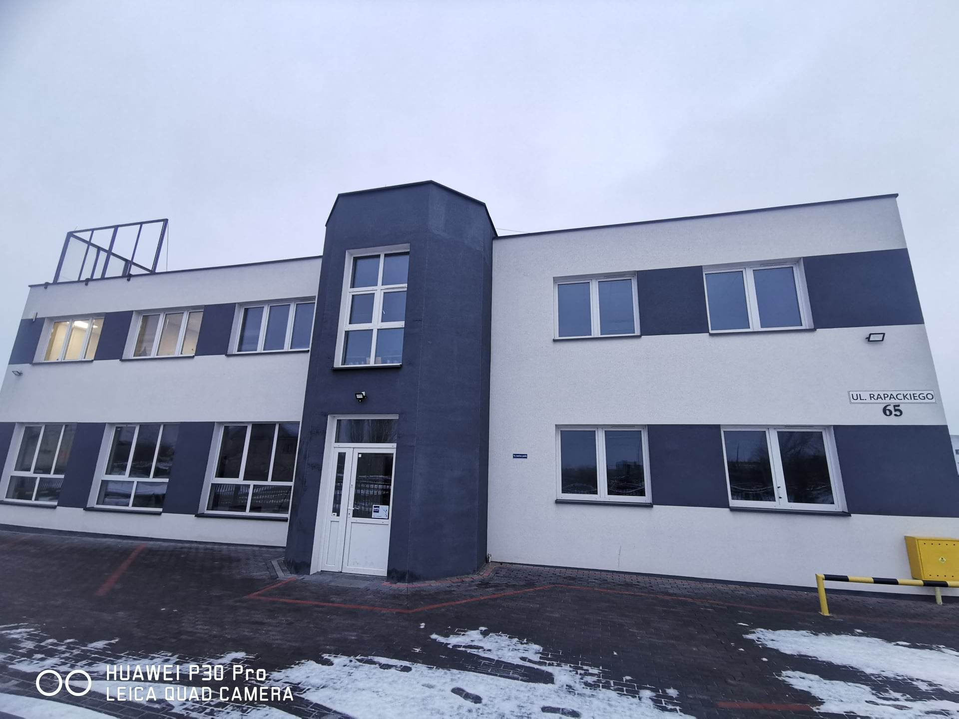 Dwukondygnacyjny budynek biurowy z szaro-białą fasadą, widoczne okna i wejście z białymi drzwiami, adres na ścianie: Ul. Rapackiego 65.