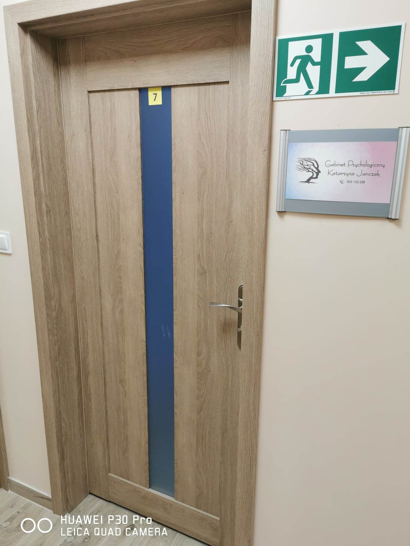 Wejście do gabinetu: drewniane drzwi z niebieskim paskiem i numerem 7, obok tabliczka informacyjna 'Gabinet Psychologiczny Katarzyna Janczak' oraz zielony znak ewakuacyjny.