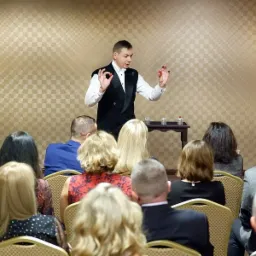 Mężczyzna w czarnej kamizelce i białej koszuli prezentuje trik z czerwonymi kulkami na tle publiczności siedzącej na krzesłach, na małym stoliku trzy szklanki i butelka wody.