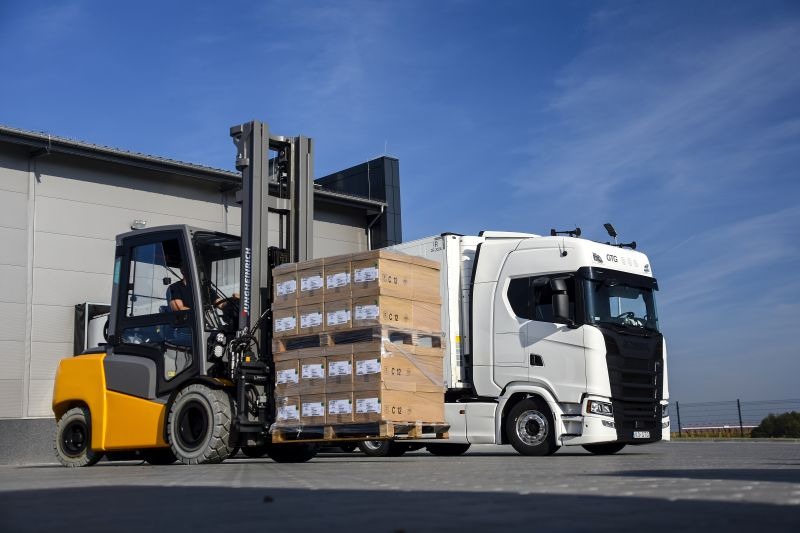Wózek widłowy Jungheinrich załadowuje palety z zapakowanymi produktami do białej ciężarówki z naczepą na tle błękitnego nieba.