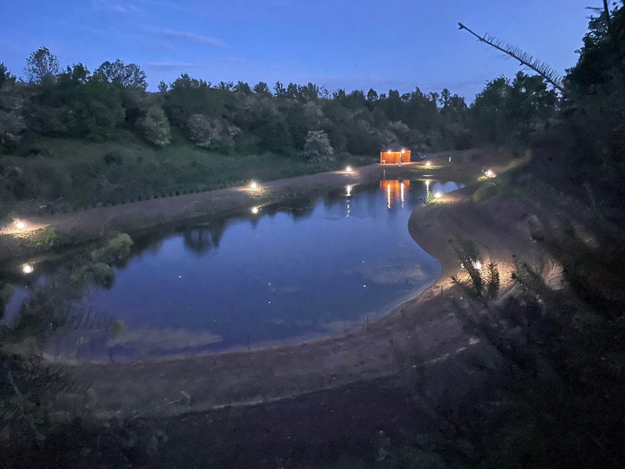 Nocny widok na wykopany staw z oświetleniem brzegowym i drewnianym domkiem na brzegu, odbijającym się w wodzie, otoczony zielenią.