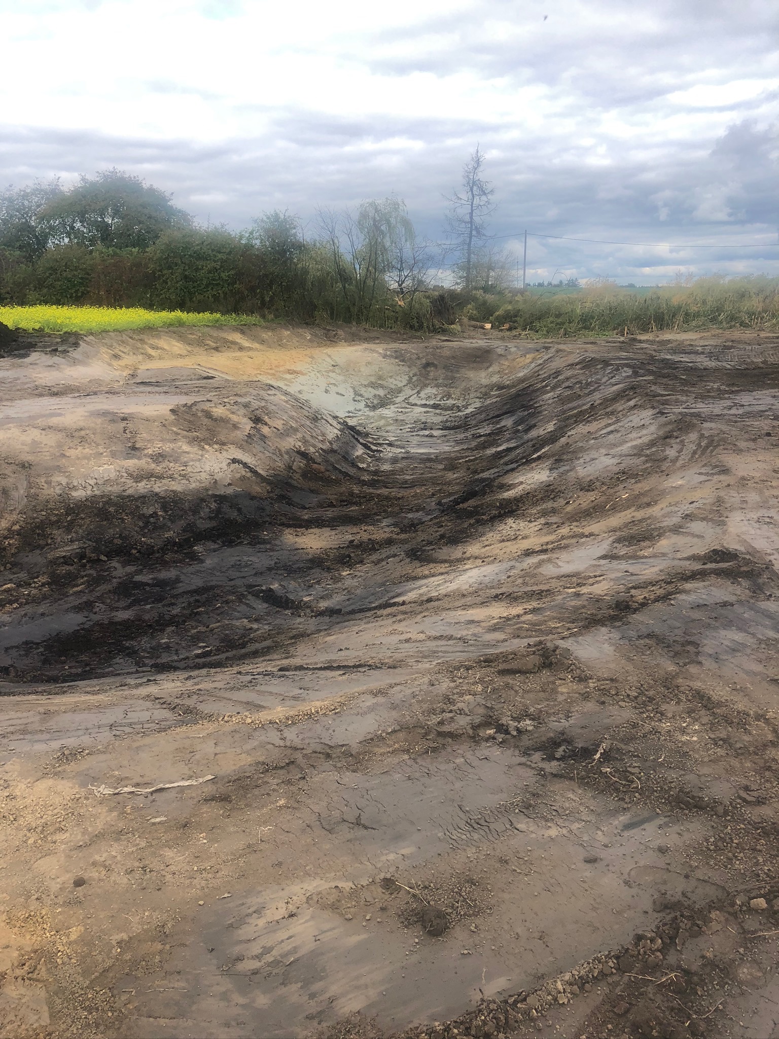 Świeżo wykopany, podłużny wykop w ziemi, otoczony roślinnością i drzewami pod pochmurnym niebem. Ziemia w wykopie jest wilgotna i ciemna, z widocznymi śladami po pracy maszyny. W tle widoczne żółte...