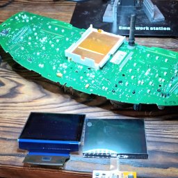 Piotr Nieznany - Rozłożony panel deski rozdzielczej samochodu z widoczną płytką drukowaną, dwoma ekranami LCD i lupą oświetleniową, na drewnianym stole warsztatowym.