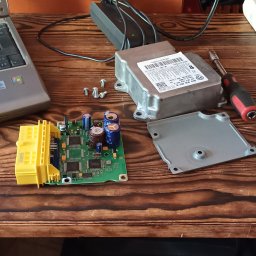 Piotr Nieznany - Otwarty sterownik samochodowy z widoczną płytką drukowaną, kondensatorami, złączem, śrubokrętem i śrubami na drewnianym stole warsztatowym, obok laptop i zasilacz.