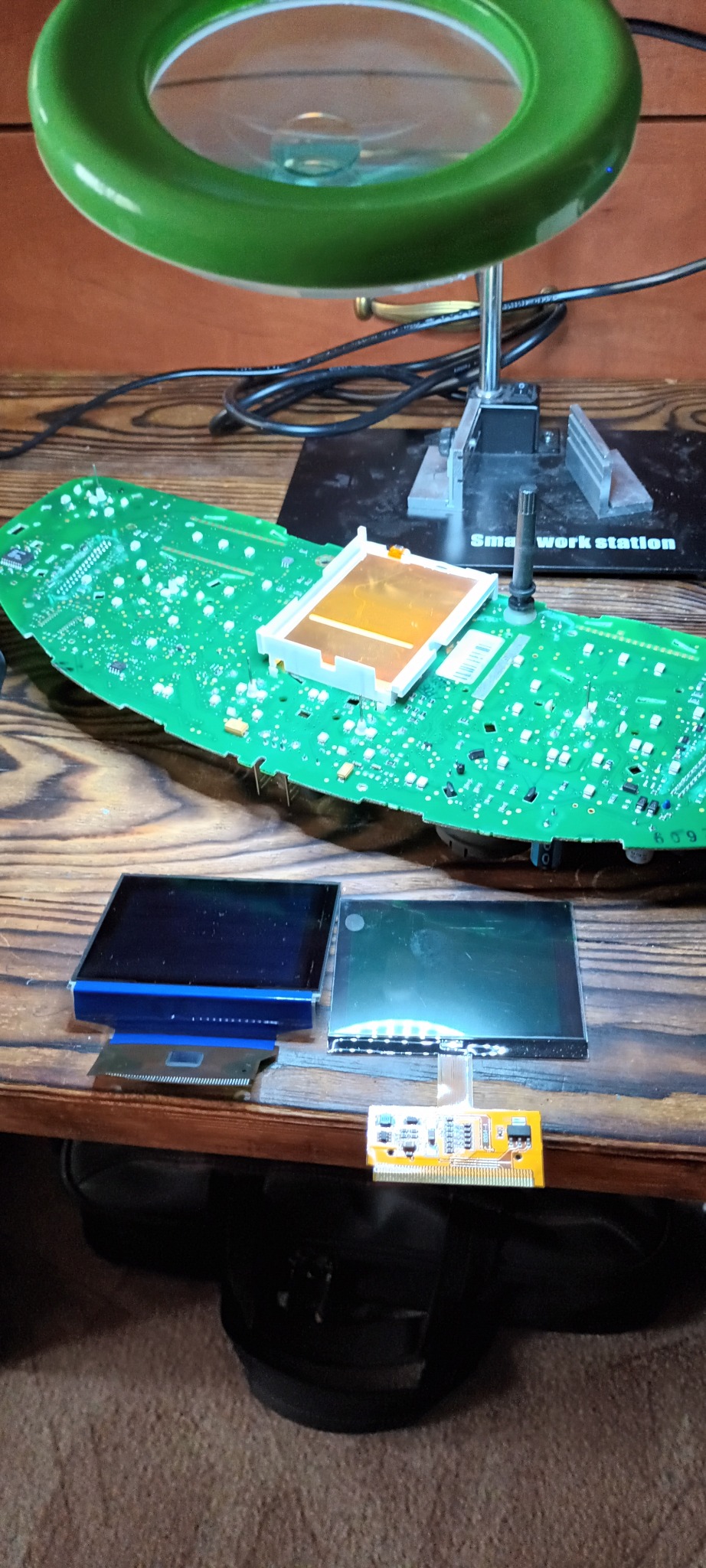 Rozłożony panel deski rozdzielczej samochodu z widoczną płytką drukowaną, dwoma ekranami LCD i lupą oświetleniową, na drewnianym stole warsztatowym.