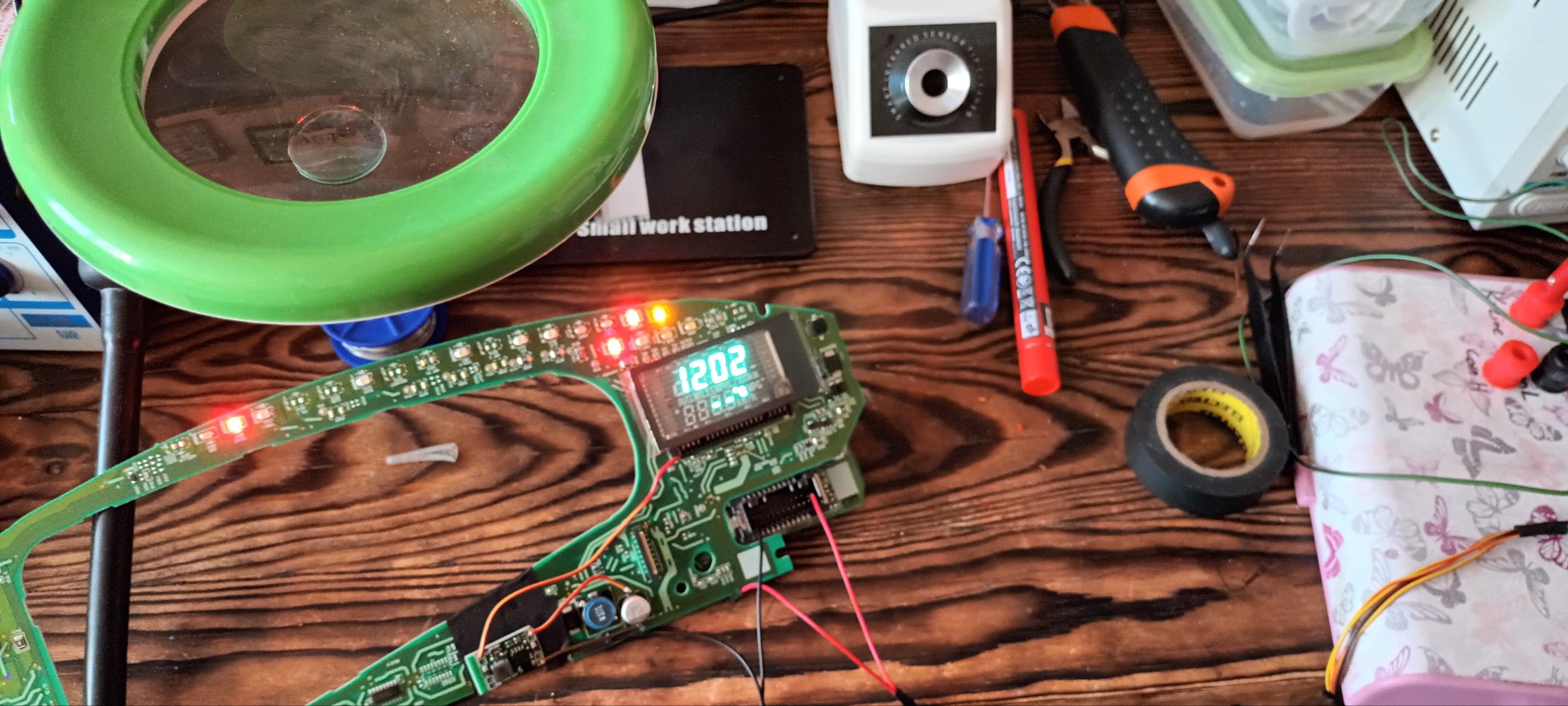 Uszkodzony panel sterowania samochodowego z wyświetlaczem, podświetlonymi diodami LED i podłączonymi przewodami, leżący na drewnianym biurku warsztatowym obok lupy, narzędzi i pudełka na drobiazgi.