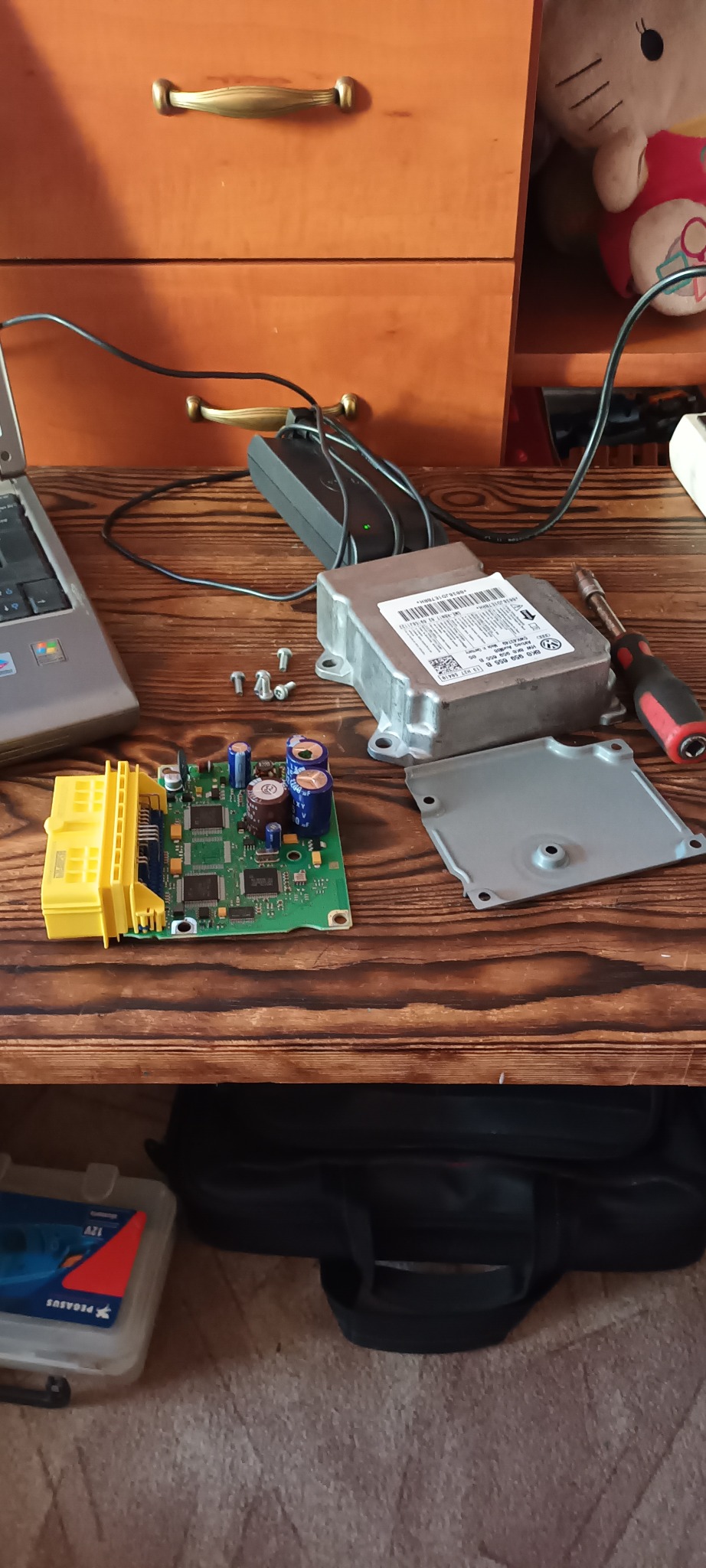 Otwarty sterownik samochodowy z widoczną płytką drukowaną, kondensatorami, złączem, śrubokrętem i śrubami na drewnianym stole warsztatowym, obok laptop i zasilacz.