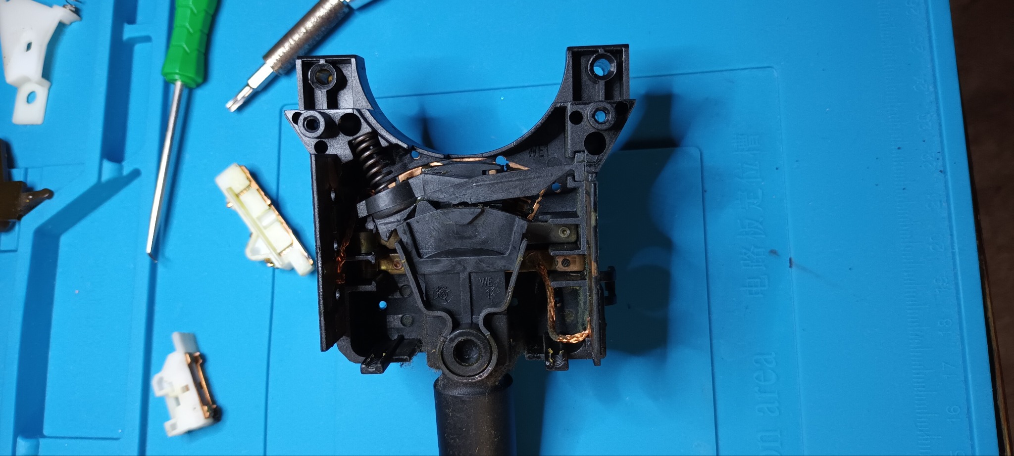 Rozmontowany przełącznik zespolony samochodu z widocznymi elementami elektrycznymi, sprężyną i śrubokrętami na niebieskiej macie warsztatowej.