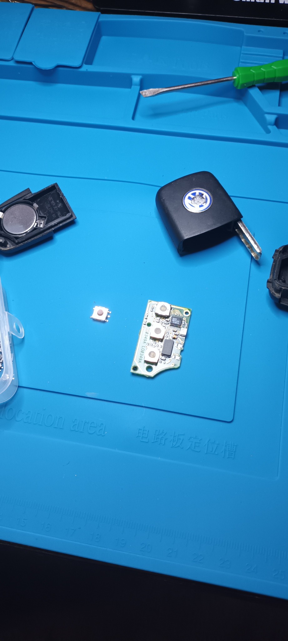 Rozłożony kluczyk samochodowy z widoczną elektroniką, baterią i obudową, leżący na niebieskiej macie warsztatowej obok małego śrubokręta.