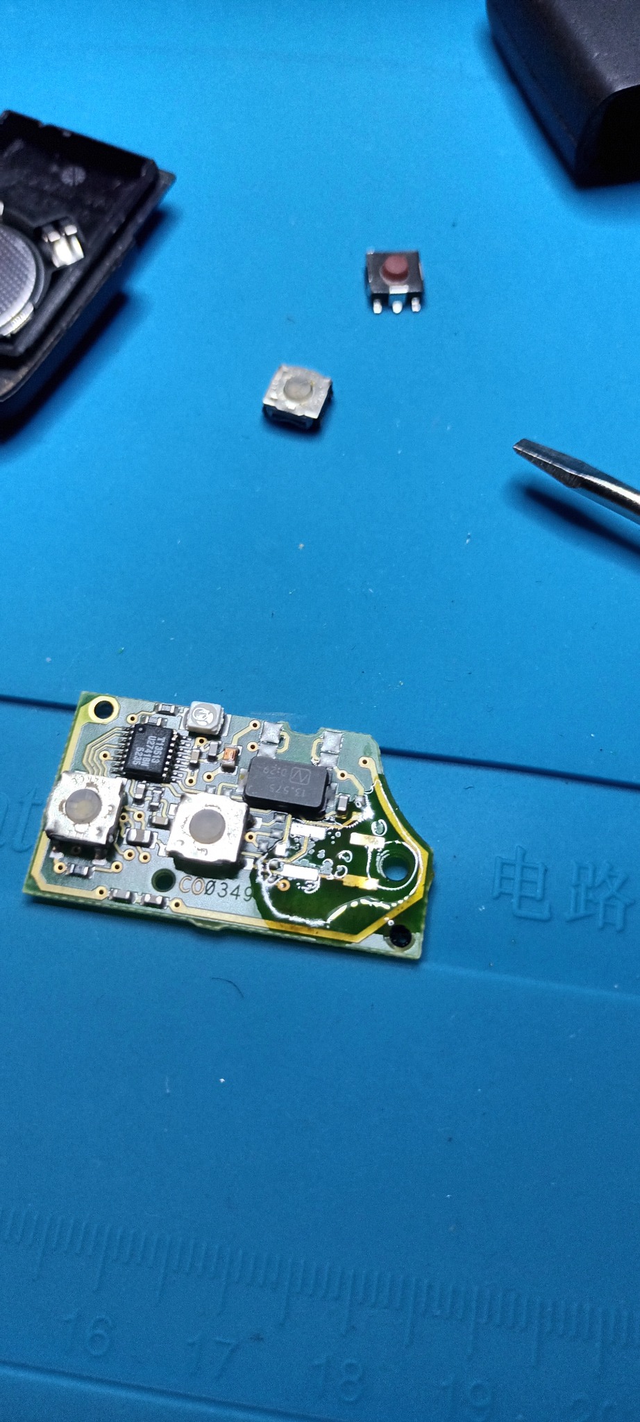 Płytka drukowana kluczyka samochodowego z widocznymi elementami elektronicznymi, przyciskami i śrubokrętem na niebieskiej macie serwisowej.