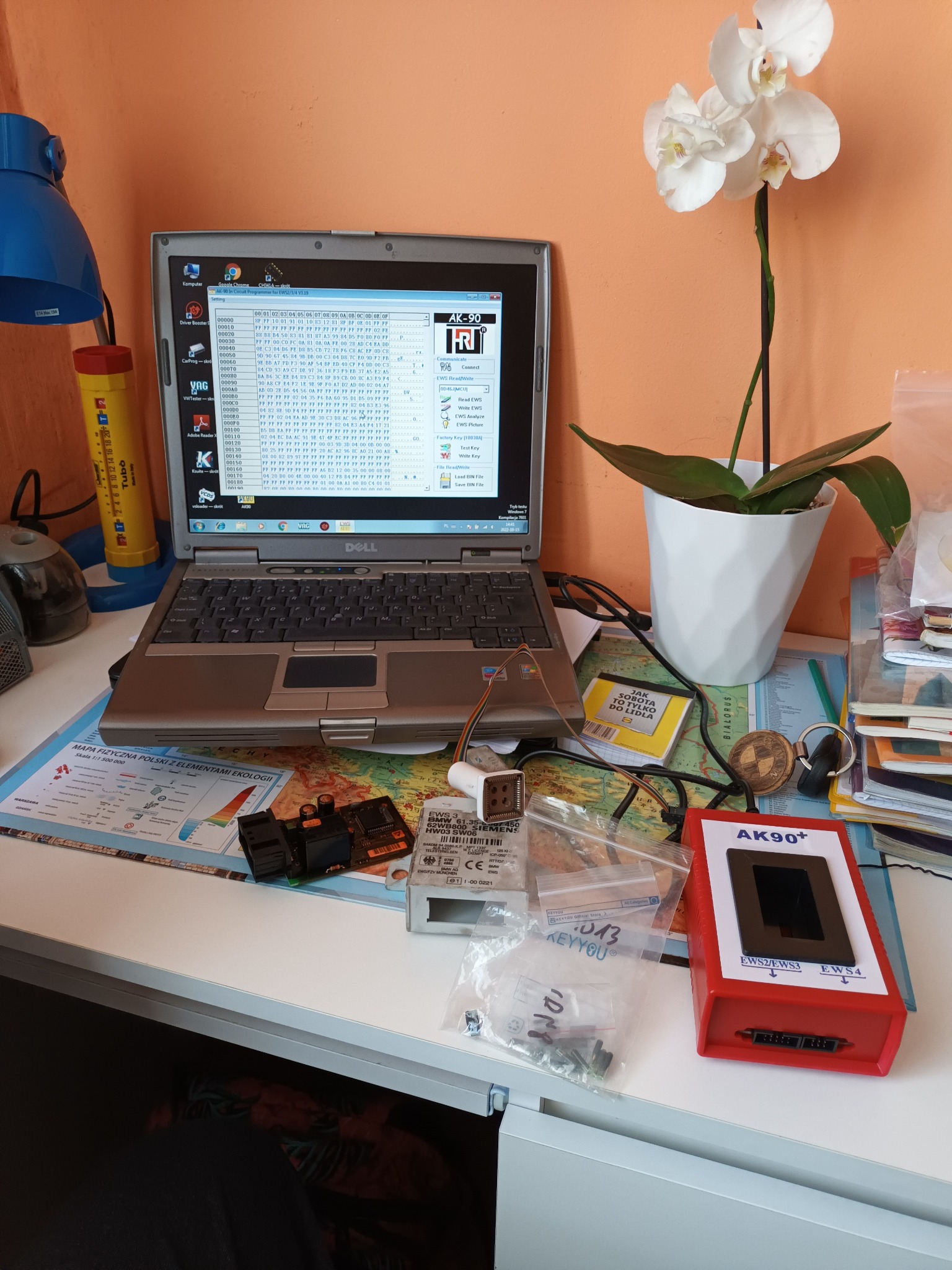 Stanowisko pracy z laptopem Dell wyświetlającym program do diagnostyki samochodowej, moduł elektroniczny, programator AK90+ i biały storczyk w tle.