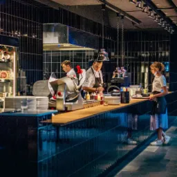 Nowoczesna kuchnia restauracyjna z dwoma kucharzami w białych uniformach przygotowującymi dania przy blacie i kelnerką w fartuchu odbierającą zamówienie, oświetlona reflektorami.