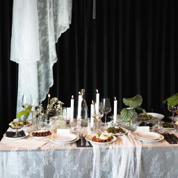 Salute - Uroczysty stół z białym obrusem koronkowym, świecami, kwiatami i elegancką zastawą na tle czarnej zasłony. Dekoracja z białej tkaniny i pereł dodaje szyku.