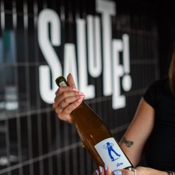 Salute - Butelka wina z etykietą 'Pepin' trzymana przez osobę z manicure i biżuterią na tle ściany z napisem 'SALUTE!'.