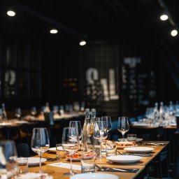 Salute - Ustawiony stół cateringowy z białymi talerzami, sztućcami, szklankami i butelkami wody na drewnianym blacie w ciemnej sali restauracyjnej z oświetleniem punktowym.