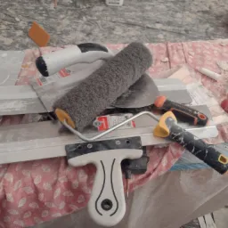 Wałek do malowania, szpachelki i inne narzędzia malarskie leżące na podłodze zabezpieczonej folią i tkaniną w trakcie prac wykończeniowych.