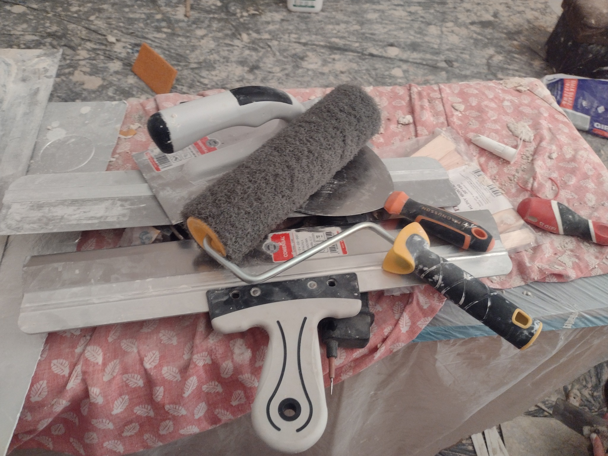 Wałek do malowania, szpachelki i inne narzędzia malarskie leżące na podłodze zabezpieczonej folią i tkaniną w trakcie prac wykończeniowych.