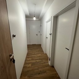 RS.Glaze Robert Skowroński - Długi korytarz w mieszkaniu z drewnianą podłogą, białymi ścianami i kilkoma białymi drzwiami, widoczna instalacja elektryczna przed montażem włącznika.