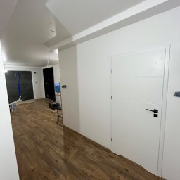 RS.Glaze Robert Skowroński - Wnętrze mieszkania po remoncie: białe ściany, drewnopodobna podłoga, białe drzwi z czarną klamką, widoczne ślady po pracach wykończeniowych w tle.