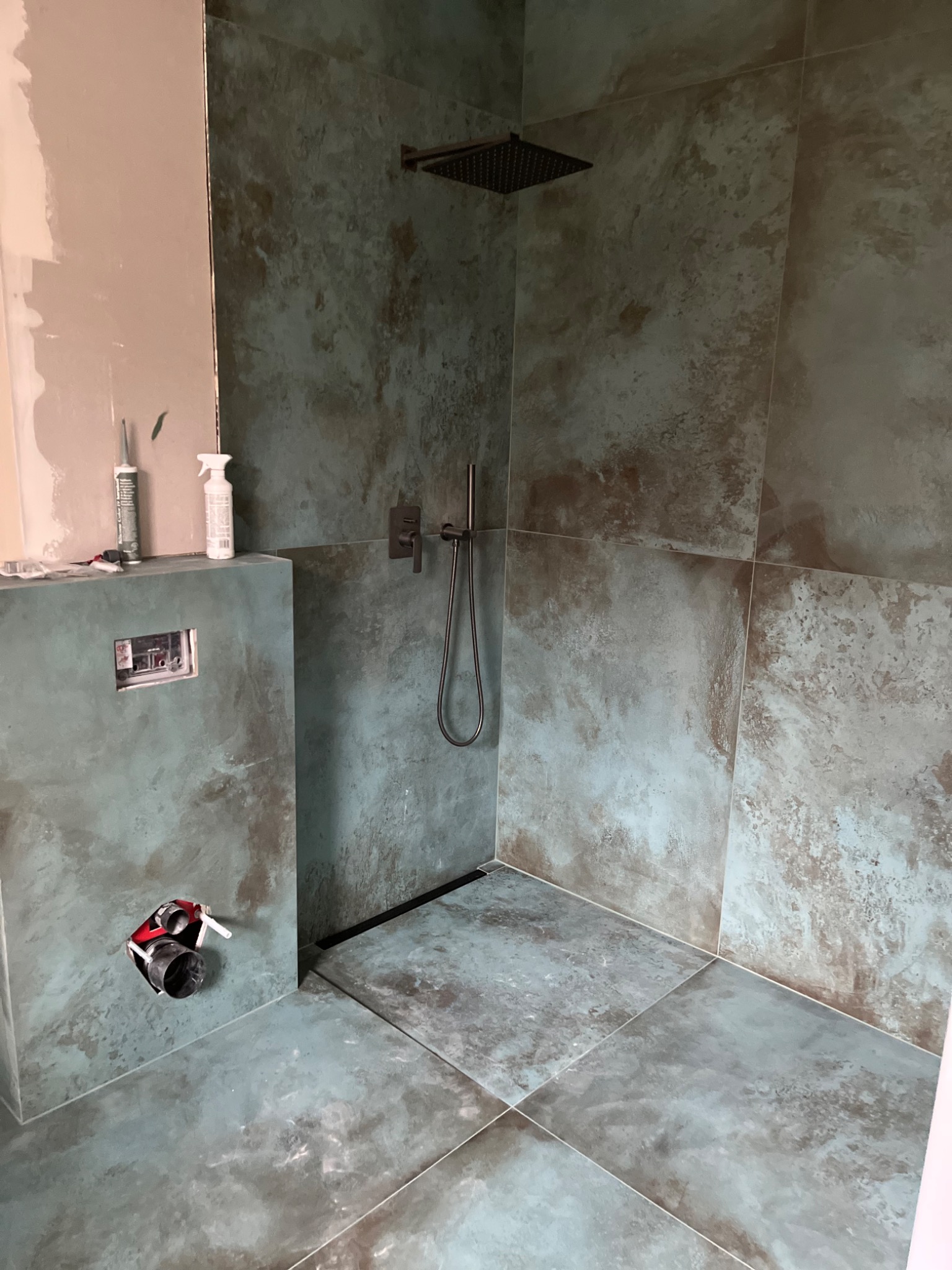 Niedokończona kabina prysznicowa z płytkami imitującymi kamień w odcieniach szarości i zieleni; widoczne narzędzia i niedokończona ściana obok.