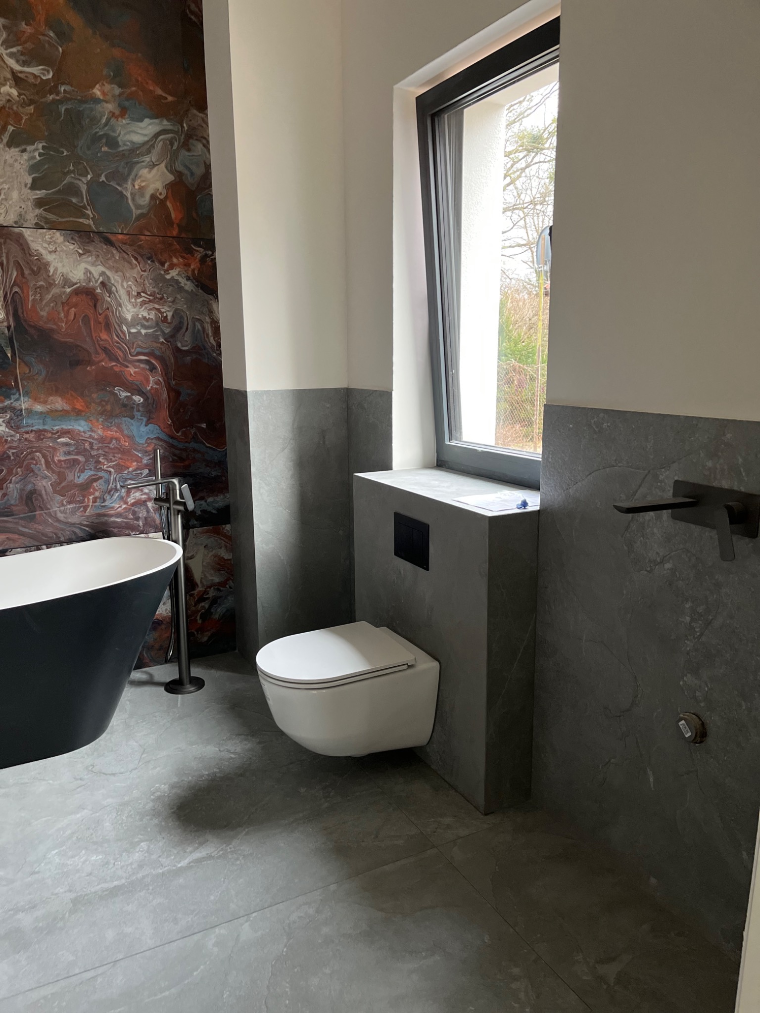 Nowoczesna łazienka z wolnostojącą wanną na tle ściany z dekoracyjnym wzorem przypominającym marmur, minimalistyczna toaleta pod oknem i szara armatura.