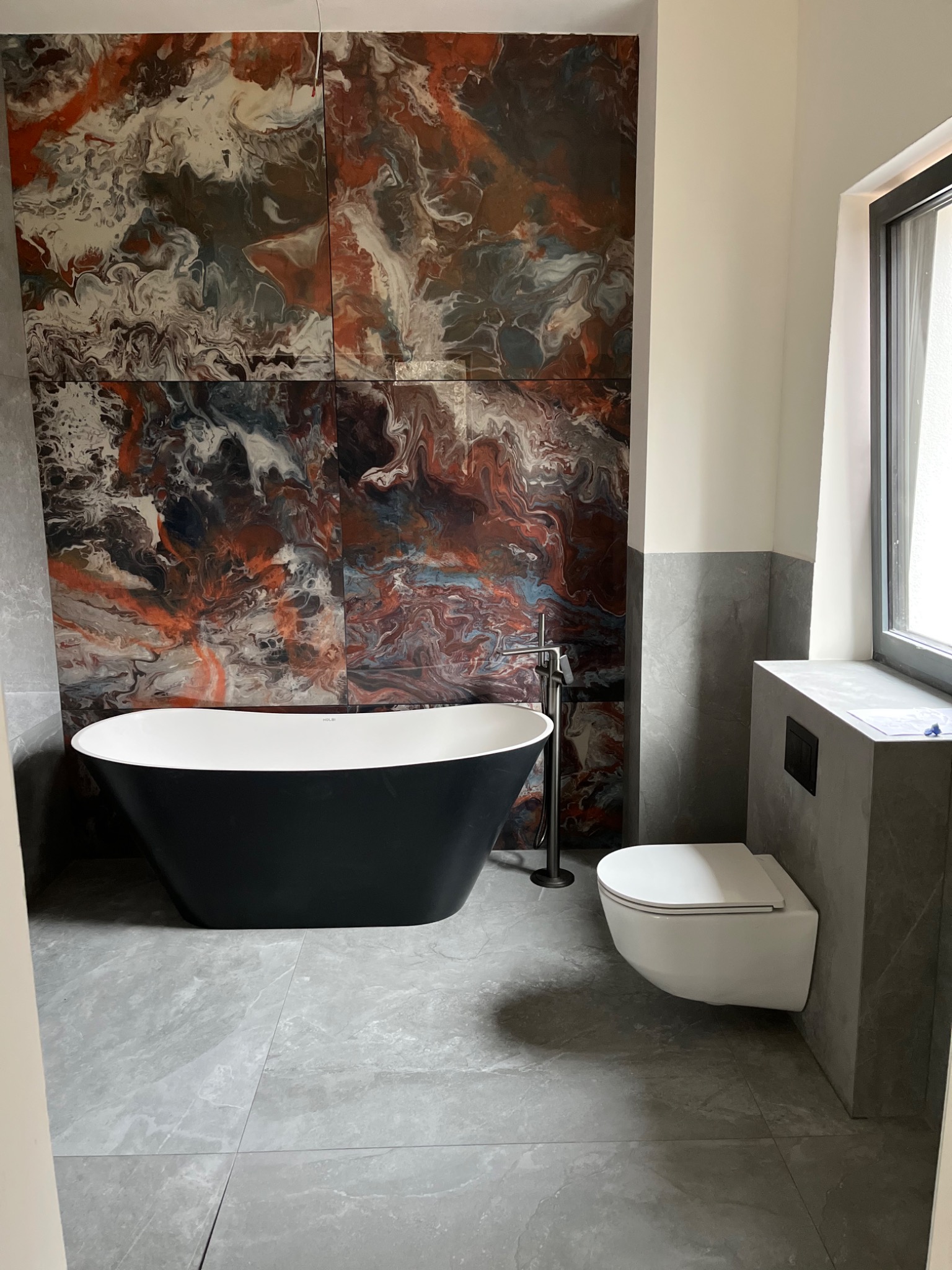 Nowoczesna łazienka z wolnostojącą czarną wanną, toaletą podwieszaną i dekoracyjnymi płytkami imitującymi marmur na ścianie.