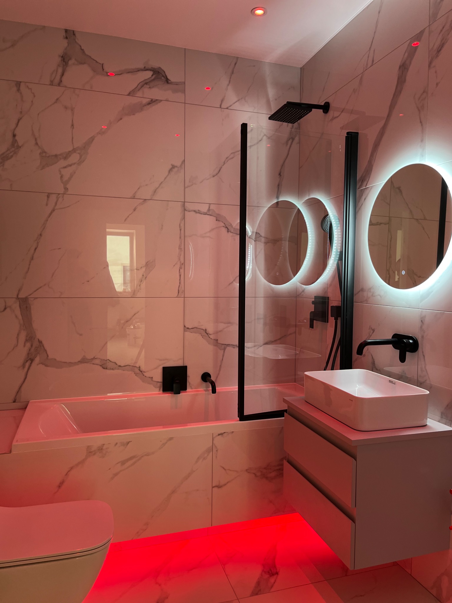 Nowoczesna łazienka z marmurowymi płytkami, podświetleniem LED w kolorze czerwonym, czarną armaturą i okrągłymi lustrami z podświetleniem.
