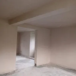 Wnętrze pomieszczenia po nałożeniu gładzi gipsowej na ściany i sufit, widoczny surowy betonowy podkład podłogowy i przejście do kolejnego pomieszczenia.