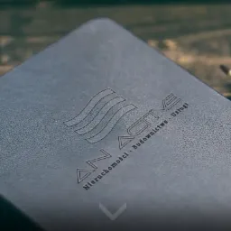 Ciemny notatnik z wytłoczonym logo 'AN ACTIVE Nieruchomości - Budownictwo - Usługi' leżący na drewnianym stole obok ołówka.