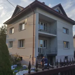 Trzykondygnacyjny dom z balkonami, świeżo po malowaniu elewacji w odcieniach szarości, widoczny pracownik schodzący po schodach i puszki z farbą w ogrodzie.