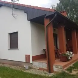 Jasny dom jednorodzinny po renowacji z nową, gładką elewacją w kolorze złamanej bieli. Widoczne brązowe rolety w oknach, czerwona dachówka oraz fragment tarasu z kolumnami. W tle zielony trawnik...