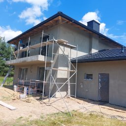 MAR-BUD Marcin Sokołowski - Dom jednorodzinny w trakcie tynkowania elewacji zewnętrznej, widoczne rusztowania, okna i drzwi zabezpieczone folią, materiały budowlane na ziemi, błękitne niebo.