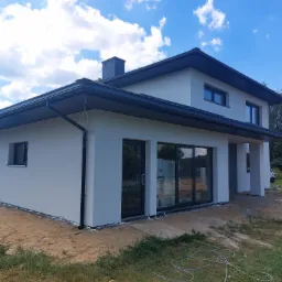 Nowoczesny dom jednorodzinny po tynkowaniu elewacji, widoczne czarne rynny i okna, teren wokół domu częściowo pokryty piaskiem.