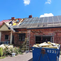 Budowa dachu domu jednorodzinnego z czerwonej cegły, widoczni robotnicy układający izolację i kontrłaty, niebieski kontener na odpady budowlane na pierwszym planie.