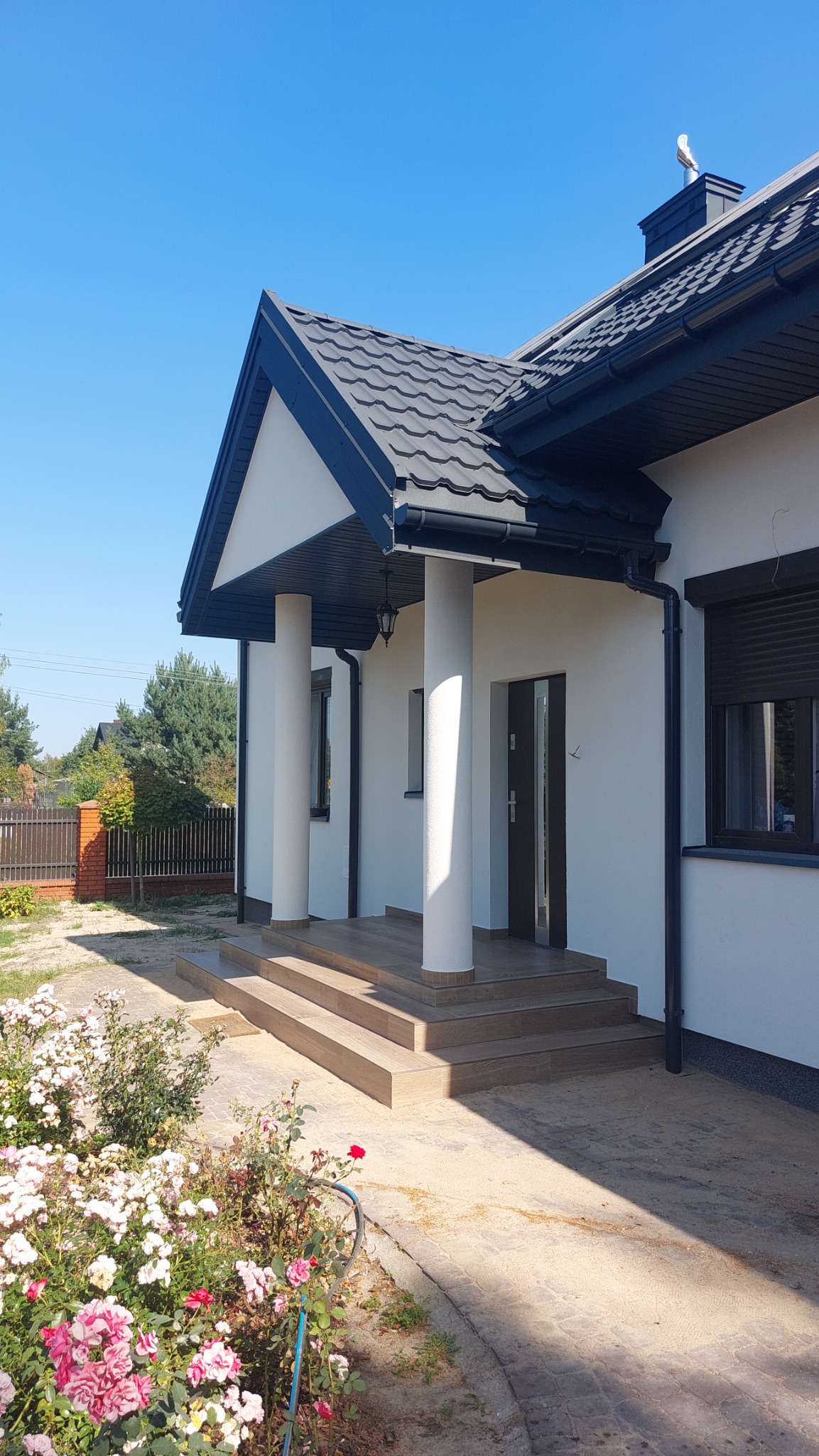 Biały dom jednorodzinny z ciemną dachówką, kolumnami przy wejściu i kwiatami w ogrodzie, widok z zewnątrz w słoneczny dzień.