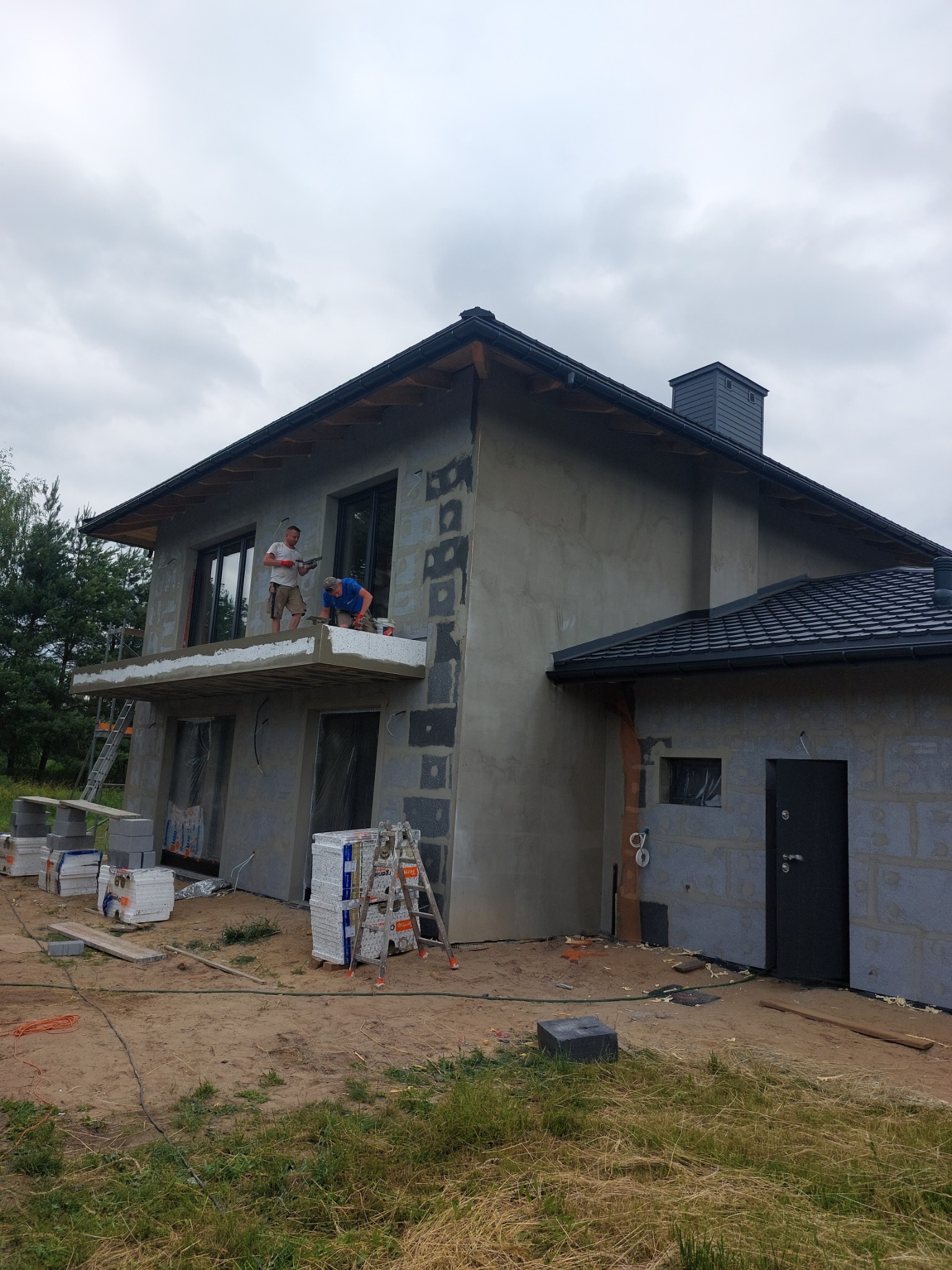 Dwóch pracowników na balkonie podczas prac wykończeniowych elewacji domu jednorodzinnego, widoczne płyty izolacyjne, otynkowana ściana i materiały budowlane na posesji.