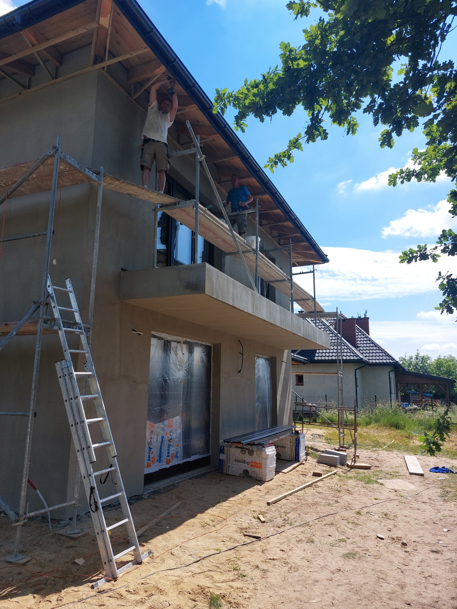 Dwóch pracowników na rusztowaniu podczas tynkowania elewacji domu jednorodzinnego; widoczne zabezpieczone folią okna i drzwi, drabina oparta o ścianę, oraz materiały budowlane obok budynku.