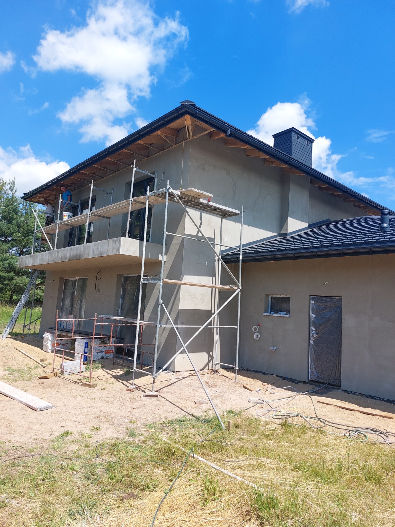 Dom jednorodzinny w trakcie tynkowania elewacji zewnętrznej, widoczne rusztowania, okna i drzwi zabezpieczone folią, materiały budowlane na ziemi, błękitne niebo.