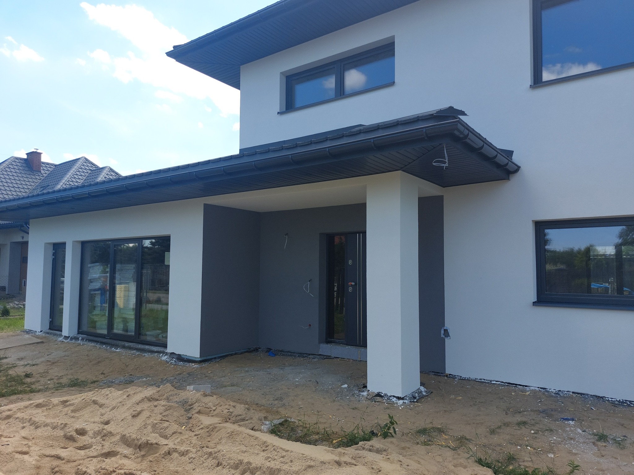 Nowoczesny dom jednorodzinny z jasnoszarą elewacją i ciemnoszarą podbitką dachową, widoczne okna z ciemnymi ramami i fragment dachu sąsiedniego budynku, teren wokół domu w stanie surowym z piaskiem.