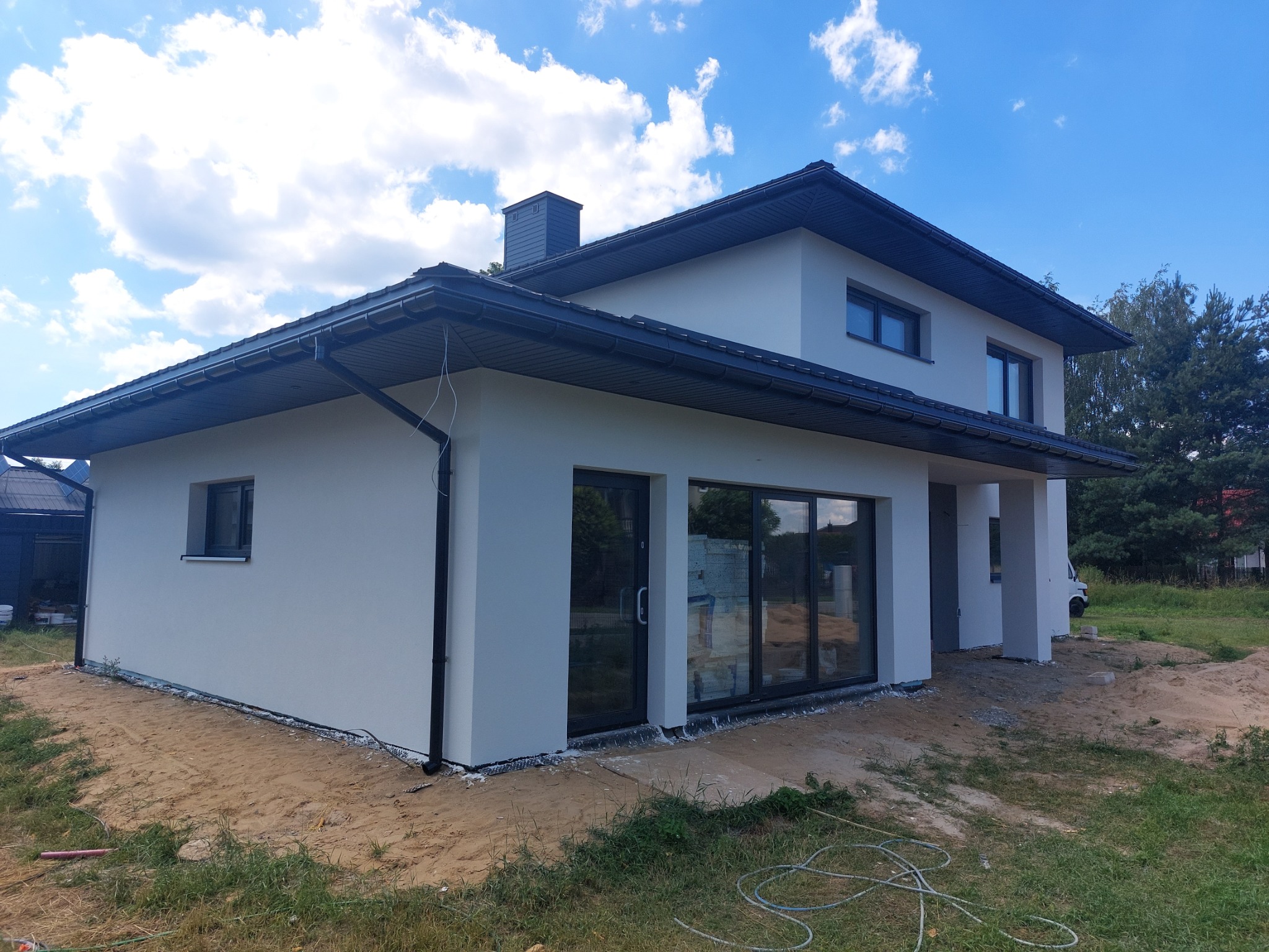 Nowoczesny dom jednorodzinny po tynkowaniu elewacji, widoczne czarne rynny i okna, teren wokół domu częściowo pokryty piaskiem.