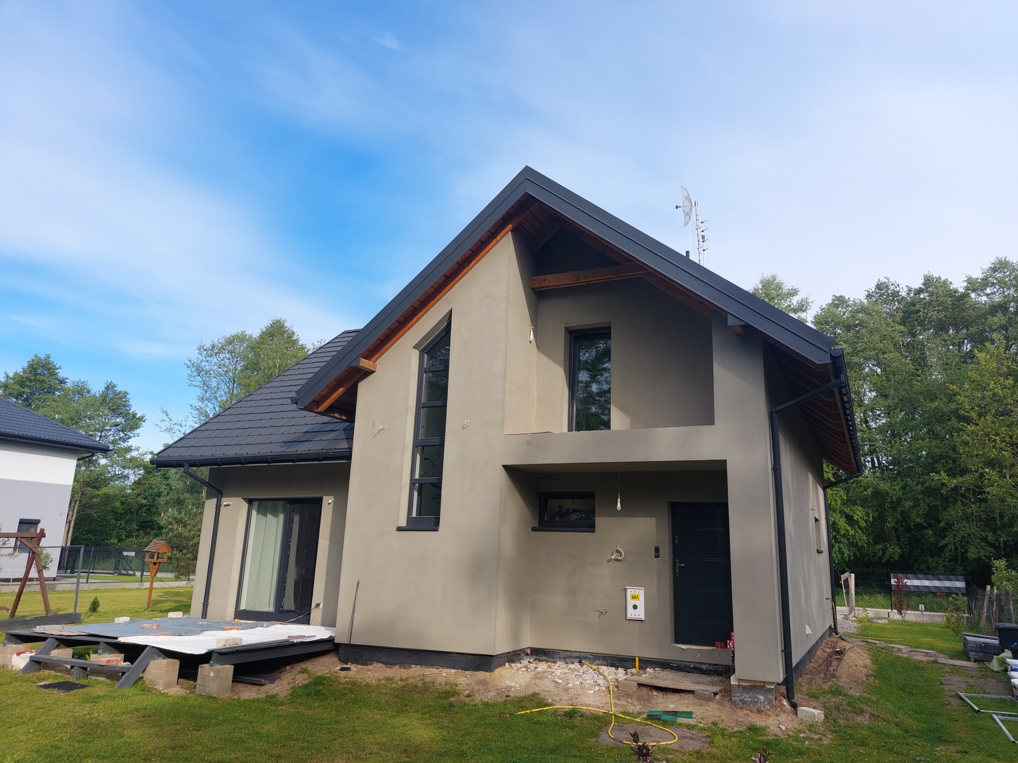 Surowa elewacja domu jednorodzinnego w trakcie prac wykończeniowych, widoczna instalacja gazowa na ścianie, przygotowane podłoże pod taras z boku budynku, w tle zieleń drzew i błękitne niebo.