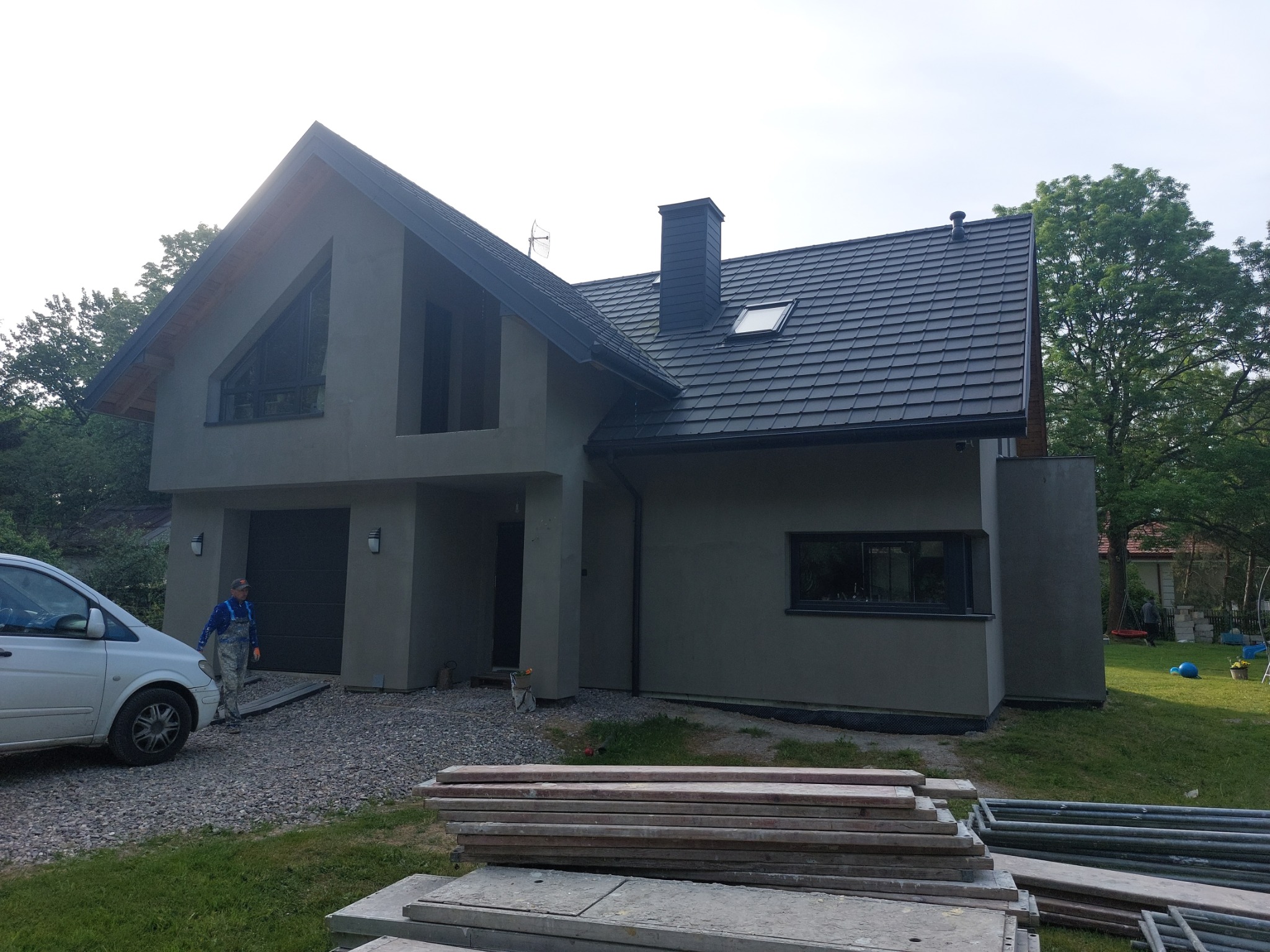 Dom jednorodzinny z szarą elewacją w trakcie prac wykończeniowych, widoczny pracownik w kombinezonie, stos desek i rusztowanie na pierwszym planie.