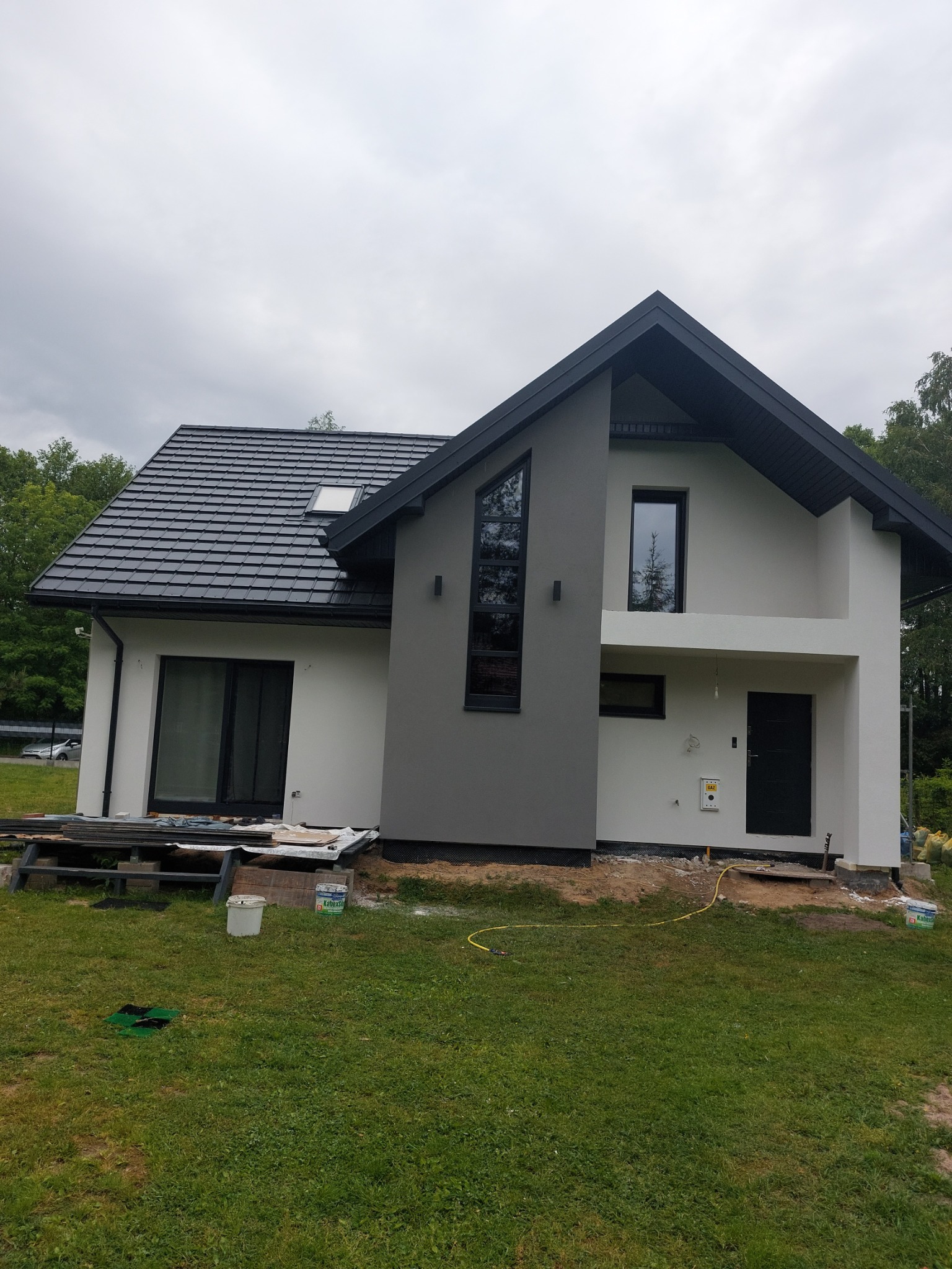 Nowoczesny dom jednorodzinny z dwukolorową elewacją (szary i biały tynk), czarną stolarką okienną i grafitową dachówką, widoczne prace wykończeniowe na zewnątrz budynku.
