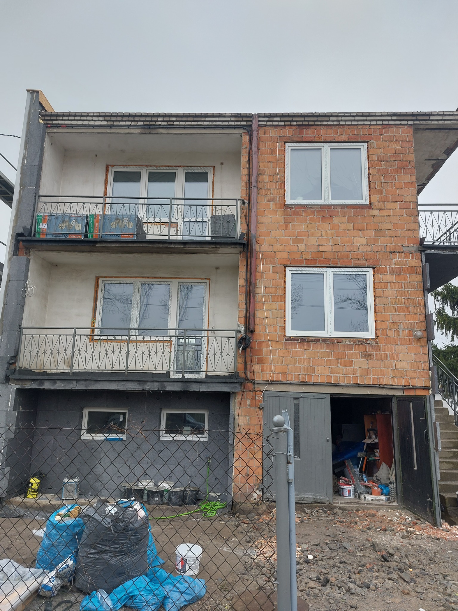 Dwupiętrowy budynek w trakcie remontu elewacji, z widoczną częścią ocieploną styropianem i częścią z surowej cegły, balkony z metalowymi barierkami, worki z odpadami budowlanymi i narzędzia...