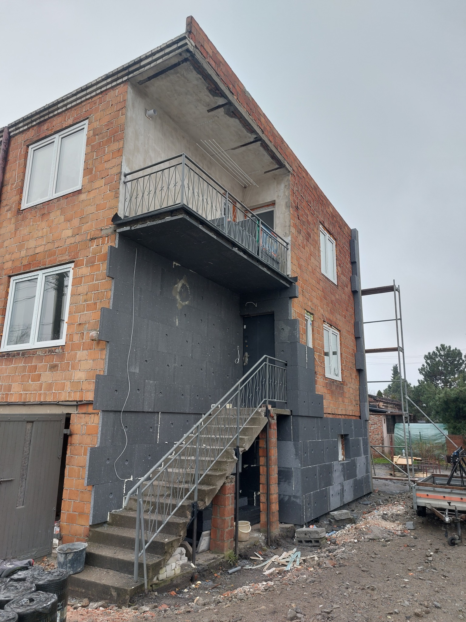 Dwupiętrowy budynek z cegły w trakcie ocieplania, widoczne szare płyty izolacyjne na fragmencie elewacji, metalowe schody wejściowe i rusztowanie z boku budynku.