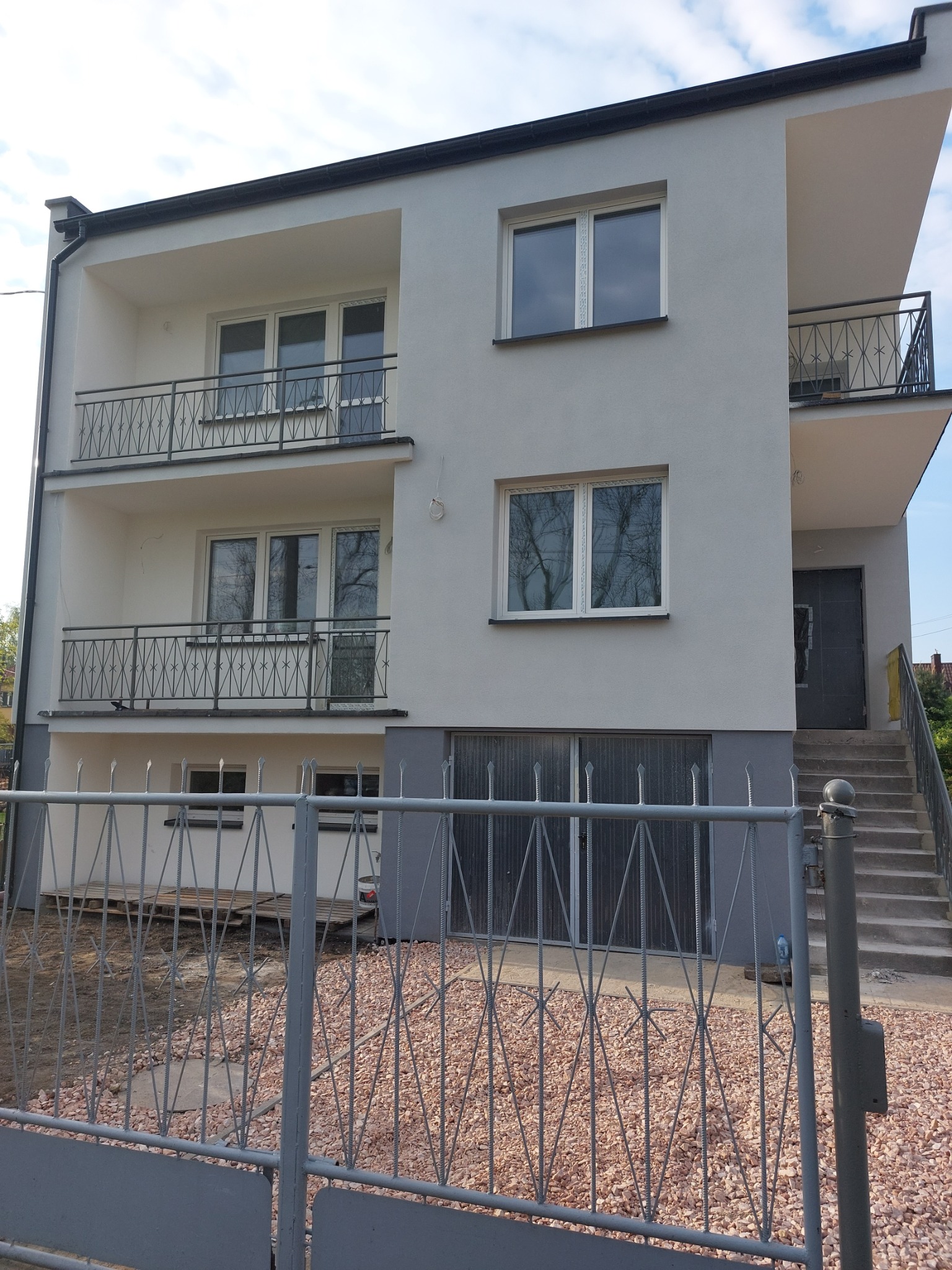Trzykondygnacyjny dom z jasnoszarą elewacją i balkonami z metalowymi balustradami, widoczny zza metalowego ogrodzenia.