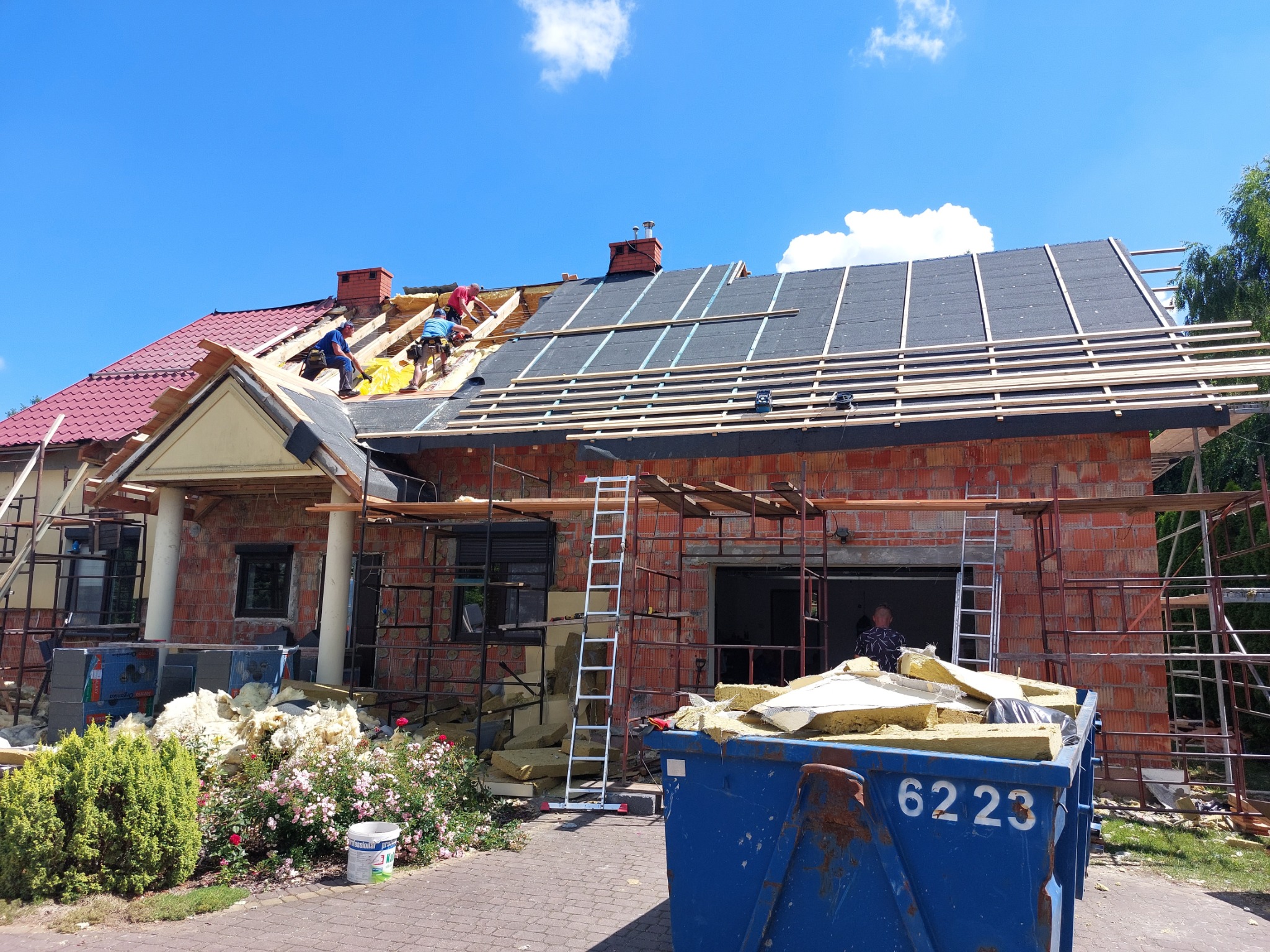 Budowa dachu domu jednorodzinnego z czerwonej cegły, widoczni robotnicy układający izolację i kontrłaty, niebieski kontener na odpady budowlane na pierwszym planie.