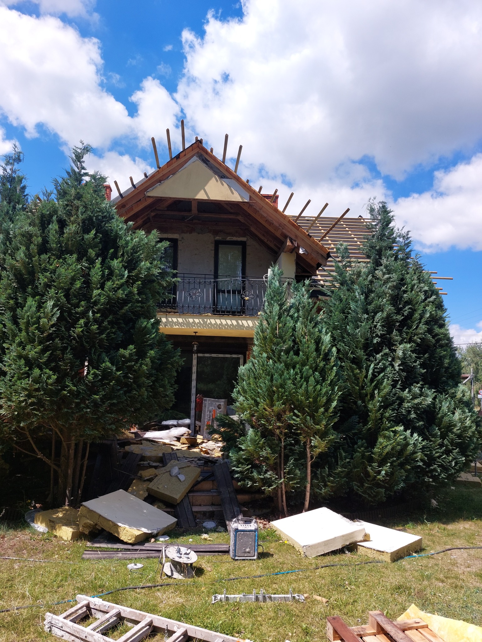 Dom w trakcie prac elewacyjnych, widoczne odsłonięte elementy konstrukcyjne dachu, fragment balkonu z balustradą, porozrzucane płyty izolacyjne i drabina na trawniku przed budynkiem, otoczony...